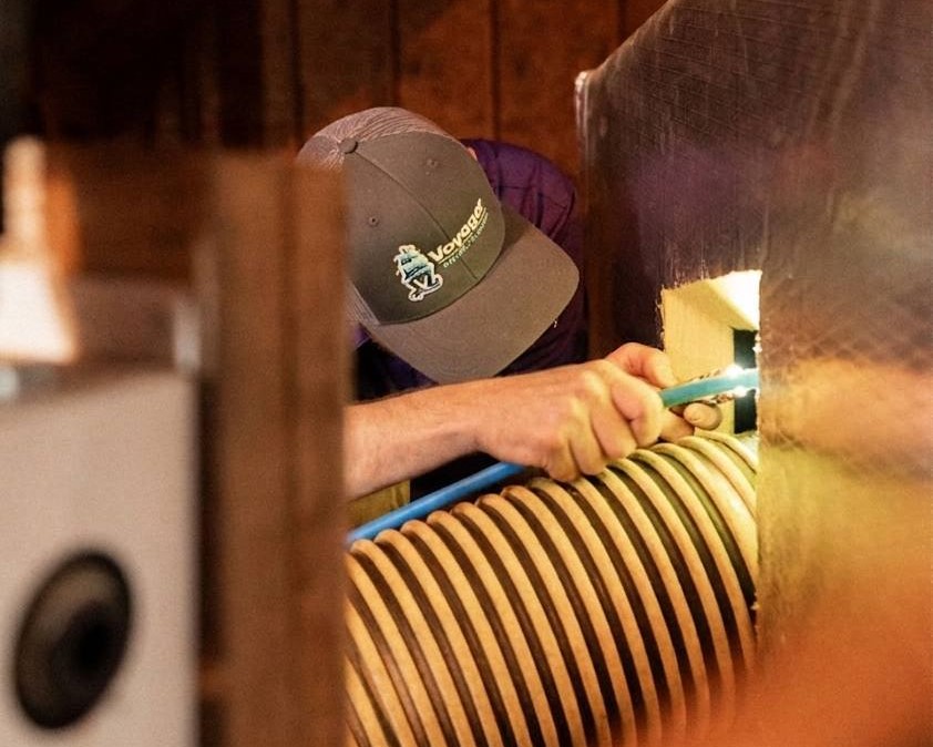 Voyager technician wearing a branded cap using a compressed air tool to clean inside a home's ductwork connected to a flexible vacuum hose.